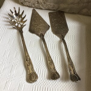May include: Three antique silver serving utensils: a slotted spoon, a pie server, and a spatula. The utensils have ornate handles with a shell design at the base. The pie server and spatula have floral patterns on their blades. The items are arranged on a white textured surface.