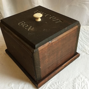 Könnte beinhalten: Eine dunkelbraune Holzkeksbox mit einem hellen Knopf auf dem Deckel. Das Wort "BISCUIT BOX" ist in den Deckel geschnitzt. Die Box hat eine rechteckige Form und ist aus Holz gefertigt.