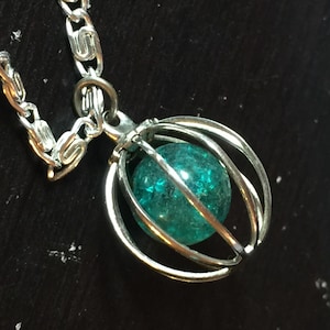 May include: A silver-toned necklace featuring a cage pendant holding a teal-colored, textured orb. The chain is made of interlocking links. The pendant is suspended against a dark background.