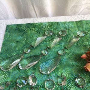 Op de afbeelding: Een verzameling van heldere kristallen prisma's, waaronder druppel-, achthoek- en rechthoekvormen, gerangschikt op een groene stoffen achtergrond.