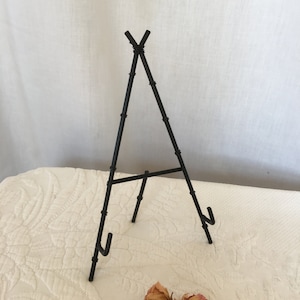 May include: Black metal easel stand for displaying photos, artwork, or other items. The easel is made of thin metal rods and has a simple, minimalist design.