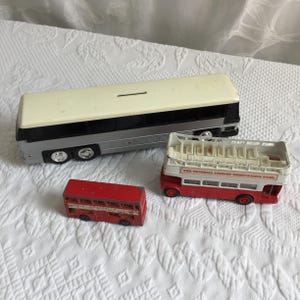 May include: Three toy buses of different sizes and designs. The largest is a silver and cream-colored bus with black accents. A red double-decker bus and a smaller red bus are also present. The buses are on a white, textured surface.