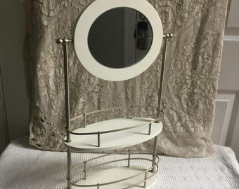 Espejo de sobremesa con repisas para maquillaje. Se puede girar para que no se vea. Blanco con metal plateado. Patas esféricas en la base.