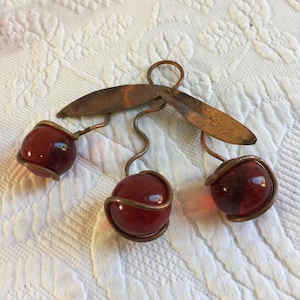 May include: A handcrafted cherry brooch featuring three red glass spheres wrapped in copper wire. The brooch has copper stems and leaves, creating a vintage, nature-inspired accessory. The brooch is a unique piece of jewelry.