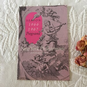 Könnte beinhalten: Ein rosa-weißes Programmheft für das New York Philharmonic Orchestra für die Saison 1966-1967. Das Cover zeigt eine Illustration einer Frau, die Geige spielt, umgeben von Cherubim.