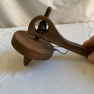 Könnte beinhalten: Ein Holz-Kreisel mit einer Schnur, die an einem Griff befestigt ist. Der Kreisel ist braun und hat eine spitze Spitze. Der Griff ist ebenfalls braun und hat ein Loch in der Mitte.
