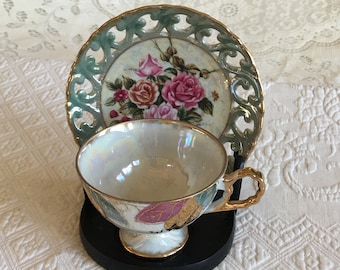 Puede incluir: Un delicado juego de taza y platillo de porcelana con un diseño floral. La taza y el platillo están decorados con rosas rosas, rojas y blancas y hojas verdes. El platillo tiene un borde festoneado y un delicado diseño calado. El juego se muestra en un soporte negro.