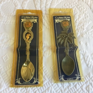 Könnte beinhalten: Zwei goldfarbene Souvenir-Löffel aus Metall in einer transparenten Kunststoff-Blisterverpackung. Der linke Löffel zeigt ein Seepferdchen-Emblem, der rechte Löffel ein Palmenmotiv. Beide Karten sind oben mit Collectible Souvenir beschriftet.