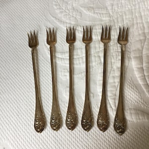 May include: Six silver-toned cocktail forks with ornate handles. The forks are arranged in a row on a white surface.