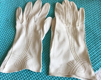 Könnte beinhalten: Ein Paar weiße Lederhandschuhe mit einem dekorativen Steppdesign. Die Handschuhe liegen auf einem türkisblauen Hintergrund.