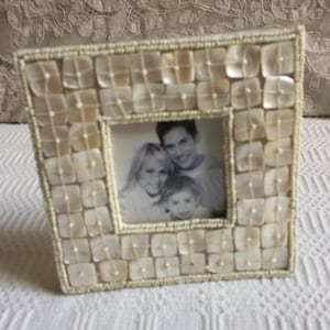 May include: A square photo frame with a cream-colored, beaded border. The frame is decorated with square, mother-of-pearl tiles. The photo inside shows a family of three. The frame is sitting on a white surface.