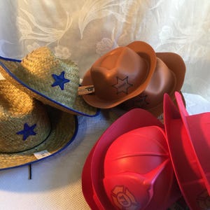May include: A collection of toy hats, including straw cowboy hats with blue stars, brown sheriff hats with a star badge, and red firefighter helmets with an FD logo.