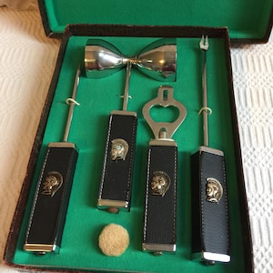 Könnte beinhalten: Vintage Bar-Werkzeug-Set in einer grün mit Samt ausgekleideten Box. Das Set enthält einen Doppel-Jigger, einen Flaschenöffner und zwei weitere Werkzeuge mit schwarzen Ledergriffen. Jeder Griff hat ein goldfarbenes Emblem. Ein kleiner, runder, beigefarbener Filzuntersetzer ist ebenfalls enthalten.