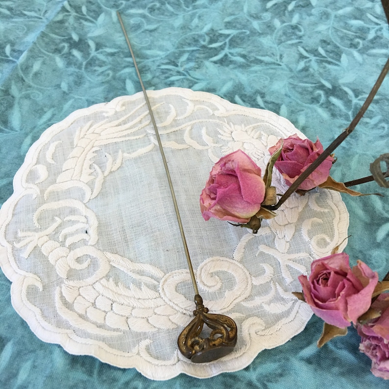 May include: A silver pin with a decorative top sits on a white lace doily with three pink roses. The doily is on a turquoise blue background.