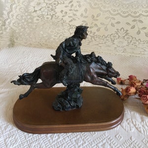 May include: A bronze sculpture of a Native American riding a horse. The sculpture is mounted on a wooden base.