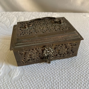 May include: A small, ornate, brass box with a hinged lid and a decorative lock. The box is intricately carved with floral patterns and has a handle on the top.