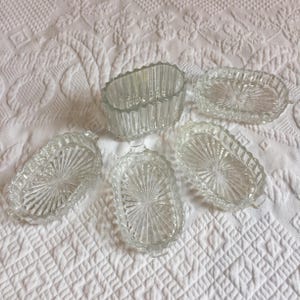 Pode incluir: Uma coleção de cinco travessas de vidro transparente. Quatro são ovais com um design de raios de sol e bordas onduladas. A quinta é retangular com ranhuras verticais. Tudo sobre uma superfície branca texturizada.