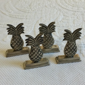 Puede incluir: Juego de cuatro porta tarjetas de mesa con forma de piña plateada. Cada porta tarjetas tiene un diseño de piña detallado y una base rectangular.