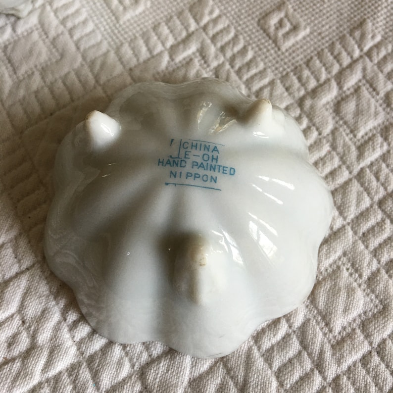 May include: White ceramic dish with a scalloped edge and three small feet. The bottom of the dish has blue text that reads "CHINA LE-OH HAND PAINTED NIPPON".