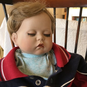 May include: A porcelain baby doll with blonde hair, wearing a blue and red striped shirt. The doll has closed eyes and a painted-on smile.