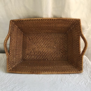May include: A rectangular woven rattan tray with curved handles. The tray is a warm brown colour and has a tightly woven pattern. The handles are integrated into the design, providing a functional and aesthetic element. The tray is suitable for serving or decorative purposes.