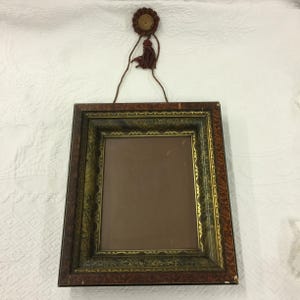 Op de afbeelding: Een vintage houten fotolijst met een versierd gouden ontwerp. De lijst heeft een bruine kwast die aan een houten knop bovenaan hangt.