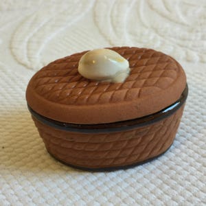 May include: A small, brown ceramic trinket box with a basket weave design. The lid has a diamond pattern and a small, white, egg-shaped knob. The box is oval-shaped and has a dark brown rim.
