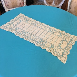 Op de afbeelding: Een witte gehaakte tafelloper met een delicaat bloemenpatroon en een rasterontwerp. De loper ligt op een turquoise tafelkleed.