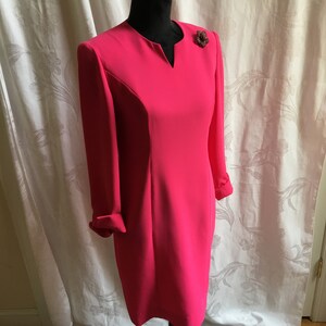 May include: A hot pink dress with a v-neck and long sleeves. The dress has a fitted silhouette and a small flower brooch on the right side of the neckline.