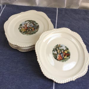 May include: A set of cream-colored, square-shaped plates with a decorative rope-like edge. Each plate features a central image of a couple in a garden setting. The plates are stacked on a blue surface.