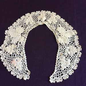 May include: A delicate white crocheted lace collar with floral motifs. The collar is made of fine thread and has a scalloped edge.