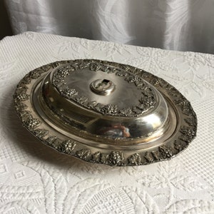 Puede incluir: Plato de servir ovalado de plata con tapa. La tapa tiene un diseño decorativo de vid y un pequeño pomo en la parte superior.