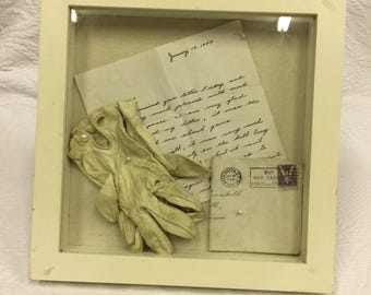 Marco de recuerdo vintage con carta y sobre de 1944 y guantes de cabritilla. Soporte de sobremesa sobre caja blanca.