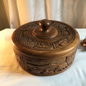 May include: A round wooden box with a carved lid. The lid features intricate designs of trees, buildings, and other natural elements. The box is made of dark brown wood and has a smooth, polished finish.