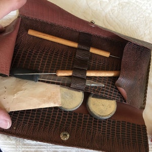 Könnte beinhalten: Geöffnete braune Ledertasche mit Reptilienhautmuster, die verschiedene Werkzeuge enthält. Enthält Werkzeuge mit Holzgriffen, ein kleines Messer und zwei runde Metallbehälter. Die Tasche wird mit einem Druckknopf verschlossen.