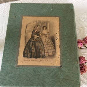 Puede incluir: Una caja verde vintage con una imagen descolorida de dos mujeres con vestidos de la década de 1850. La imagen está titulada "Godefroy's Universal Colored Fashions 1850".