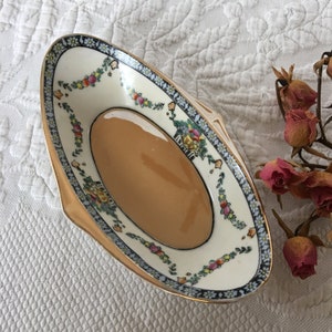May include: An oval-shaped ceramic dish with a gold rim and a floral design. The dish is decorated with a light brown glaze and features a white background with colorful flowers and leaves.