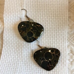 May include: A pair of earrings with a dark green and gold pattern. The earrings are teardrop-shaped with a hook closure. The pattern has a speckled design with gold accents. The earrings are displayed on a white textured surface.