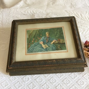 Puede incluir: Un marco de fotos de madera vintage con un acabado dorado. El marco contiene una impresión de una mujer con un vestido azul sentada en un entorno de jardín. La impresión está titulada "MADAME DE POMPADOUR".