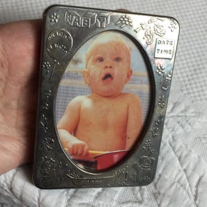 Könnte beinhalten: Silberfarbener Baby-Fotorahmen aus Metall mit ovaler Öffnung. Der Rahmen ist mit floralen und Baby-Themen-Verzierungen verziert, einschließlich des Wortes "BABY" in Blockbuchstaben. Der Rahmen enthält auch die Wörter "WEIGHT" und "LENGTH".