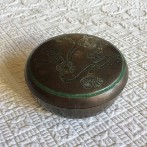 Peut inclure: Une boîte à bijoux ronde en bronze avec un bord vert et un motif floral. La boîte a un aspect légèrement usé.