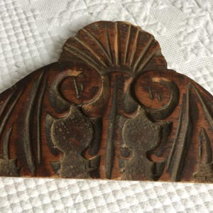 Può includere: Un intaglio in legno semicircolare caratterizzato da un motivo a raggiera nella parte centrale superiore, affiancato da due figure simmetriche stilizzate con forme curve simili a corna, su uno sfondo con venature del legno naturale scuro.