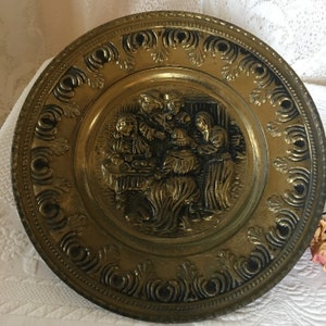 Vintage Gold Tin Ornate Plate With Wall Hanger Ring on Back. Pioneer People At Table in the Center With Swirling Designs Around Edge.