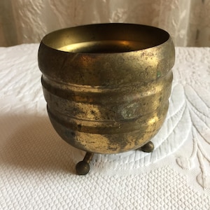 May include: A small, brass planter with a ribbed design and three legs. The planter has a worn, aged look.