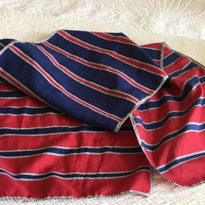 May include: A folded scarf with a striped pattern in red, navy blue, and silver. The scarf is made of a soft fabric and has a scalloped edge. The stripes run horizontally across the scarf.