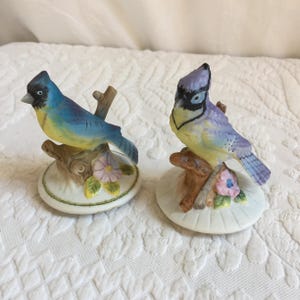 Könnte beinhalten: Zwei Porzellan-Blauvogel-Figuren, die auf Baumästen mit Blumen sitzen. Die Vögel sind mit gelben, blauen und violetten Akzenten bemalt.