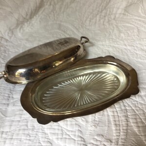 May include: A silver-colored butter dish with a lid. The dish has an oval shape with a decorative, scalloped edge. The lid has a handle and is slightly ajar, revealing the interior of the dish.