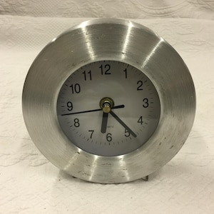 Puede incluir: Un reloj de cuarzo redondo de metal cepillado con esfera blanca y números negros. El reloj tiene manecillas negras y un centro dorado. La hora es aproximadamente las 6:26. El reloj está sobre una superficie blanca.