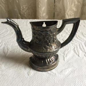 Puede incluir: Tetera de plata con un diseño ornamentado, que presenta un patrón floral y un pico con un asa curva. La tetera tiene un agujero en la parte posterior para colgarla.