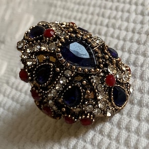May include: An ornate, antique-style brooch with a gold-tone metal base. The brooch is encrusted with various gemstones, including large, teardrop-shaped dark blue stones, small red stones, and clear crystals.